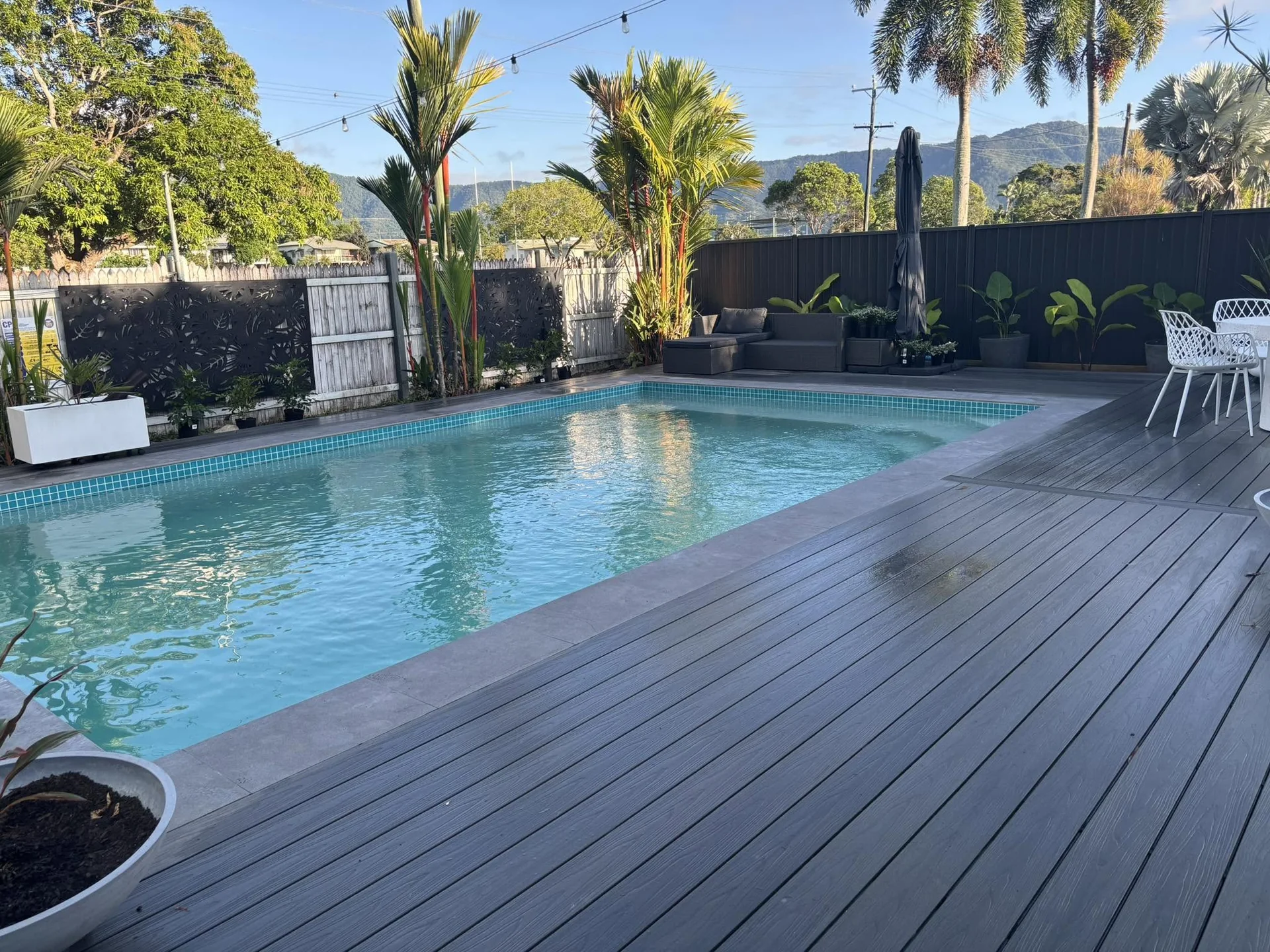 Finished rectangular swimming pool with grey decking, tropical planting and mountain backdrop.